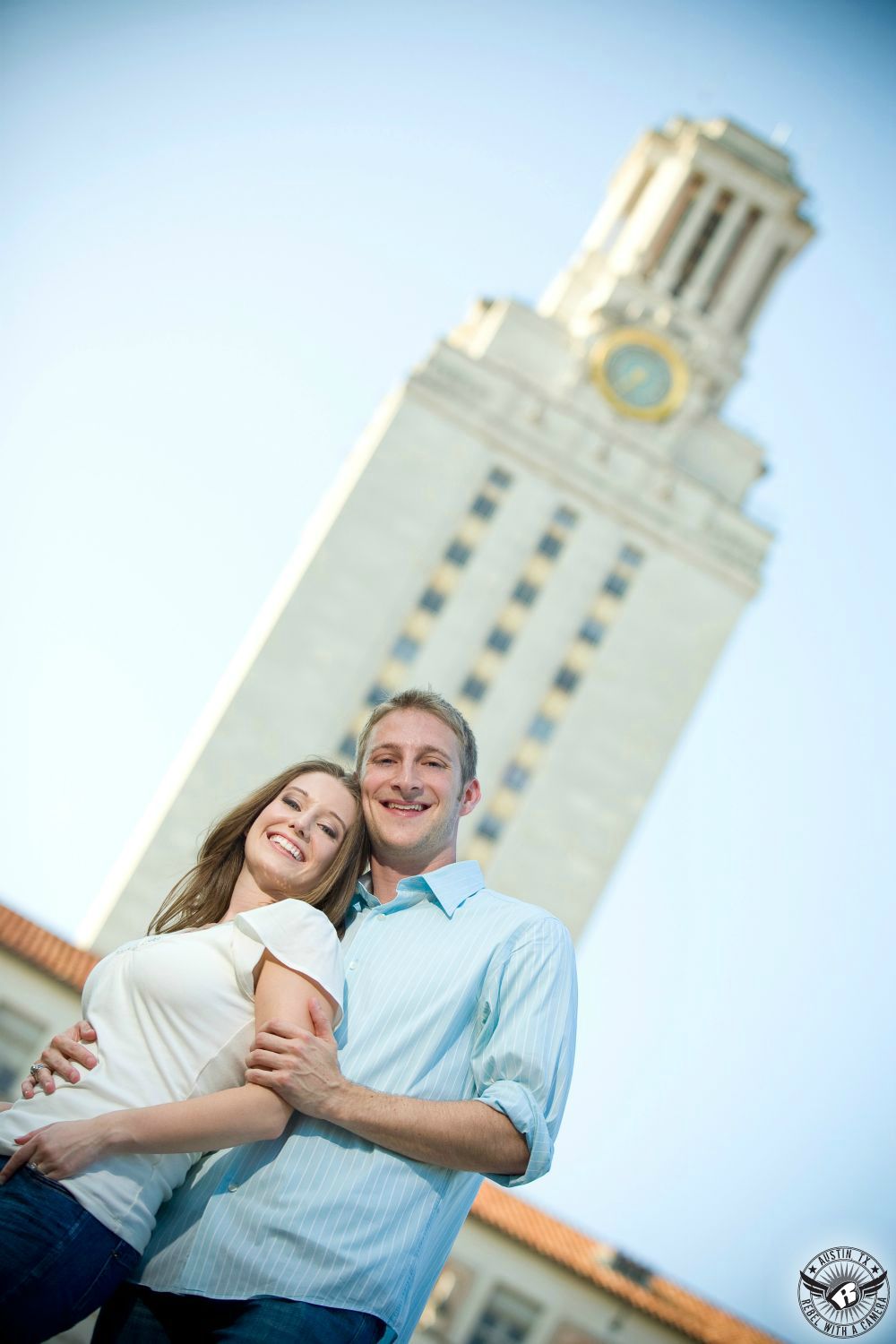 happy sandy haired girl wearing a white blouse and blue jeans is embraced by a happy sandy haired guy  with a blue button up shirt in front of the clock tower at UT Austin in this engagement portrait in texas